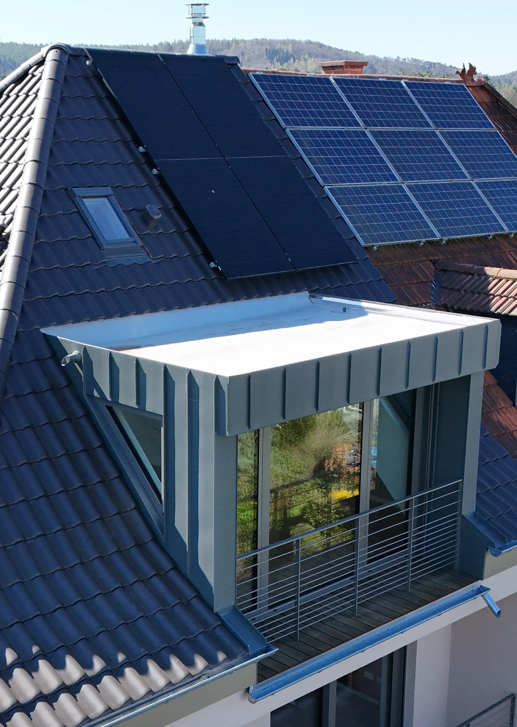 Kuhn & Mazeres – Dachdeckerei – Dunkel eingedecktes Wohnhausdach mit einer modernen, zurückgesetzten Gaube (Loggia-Gaube), ausgestattet mit kleinem Balkon und Stehfalzverkleidung aus Metall sowie integrierten Solarthermie- und Photovoltaikmodulen.