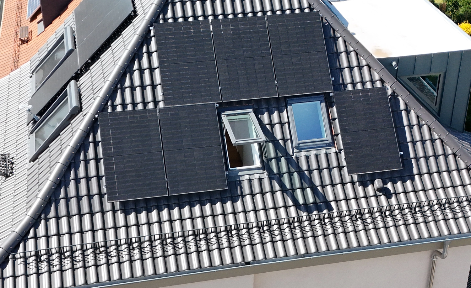 Kuhn & Mazeres – Dachdeckerei – Dunkel eingedecktes Wohnhausdach mit einer Photovoltaikanlage (PV), deren Module um die Dachfenster herum angeordnet sind. Monokristalline „All-Black“-Solarmodule sind sauber auf der geneigten Dachfläche (Ziegeldeckung) neben einem geöffneten Dachfenster montiert